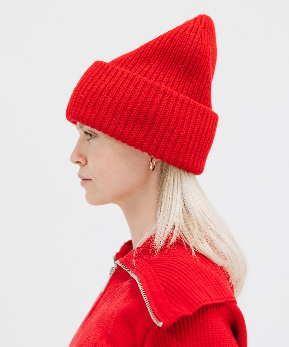 A woman wearing a ruby red colored ribbed beanie and a red jacket facing sideways against a plain background #color_ruby red