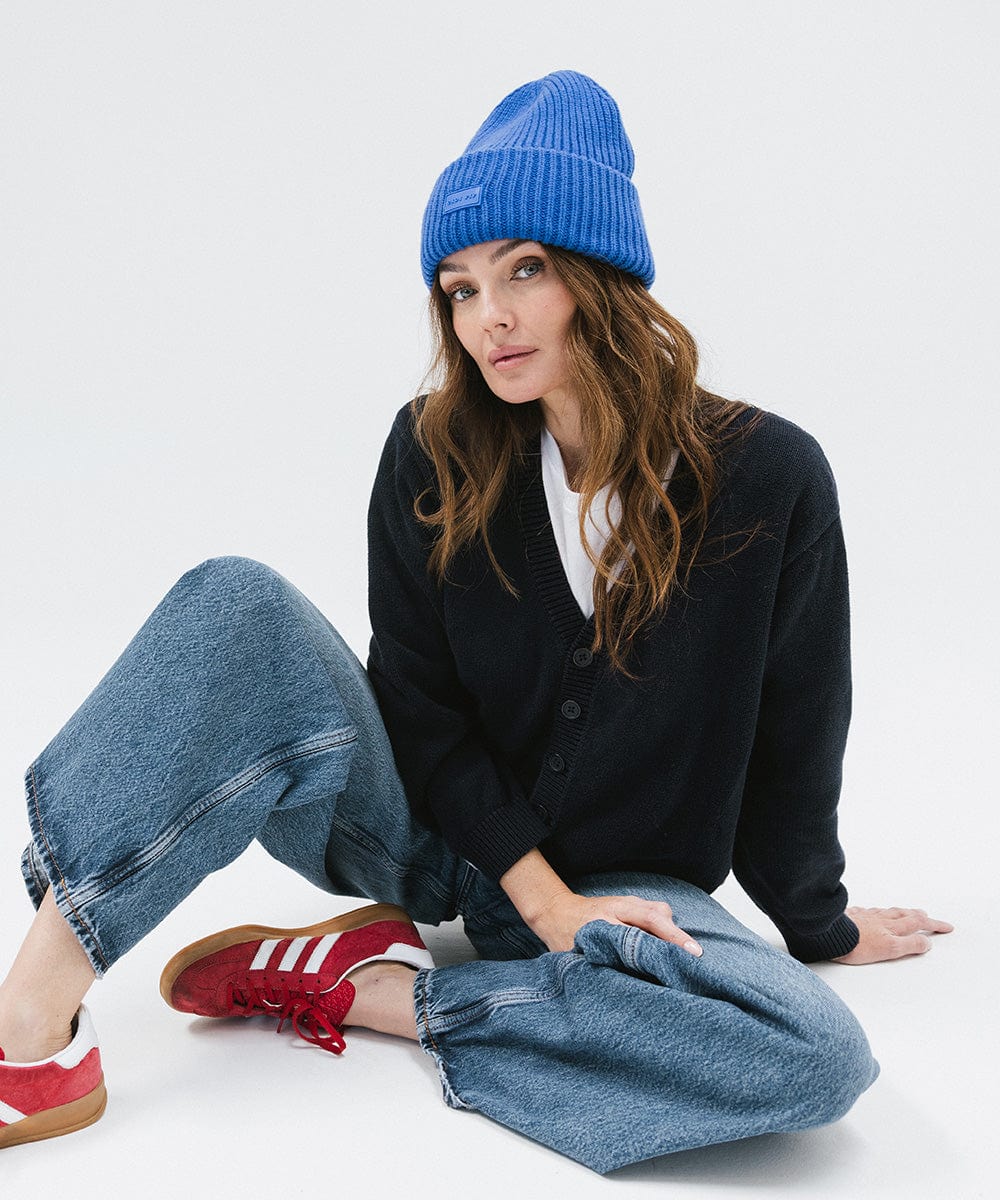 A woman wearing an alpine blue colored ribbed beanie, a dark blue sweater, blue jeans, and red shoes, sitting against a plain background #color_alpine blue
