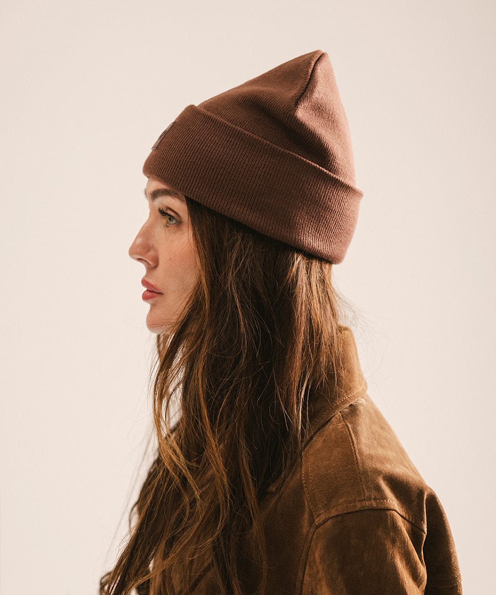 Woman wearing a chocolate colored beanie and brown jacket, facing sideways against a plain background #color_chocolate