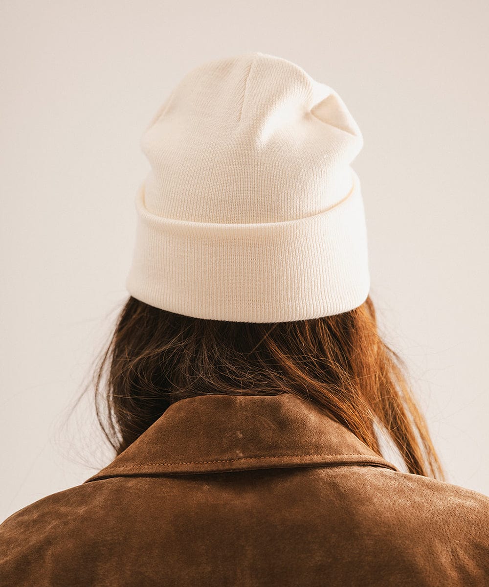 Person wearing an off white beanie and brown jacket, facing behind against a plain background #color_off white