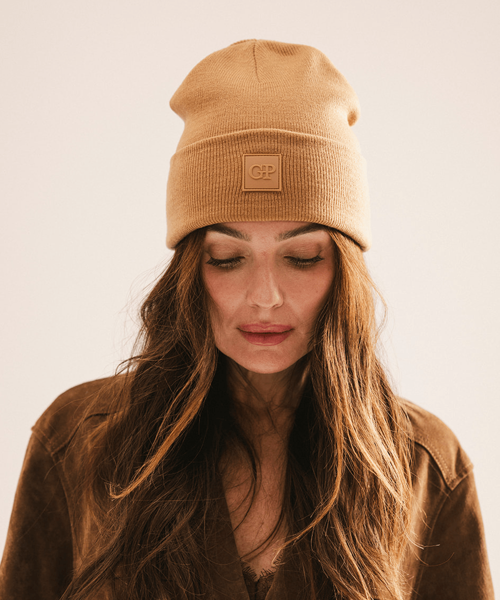 Woman wearing a latte colored beanie with a brand logo, looking down against a plain background #color_latte