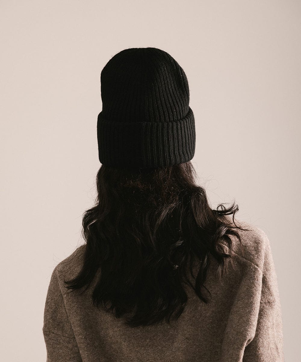 Person wearing a black knit beanie and brown coat on a beige background #color_black