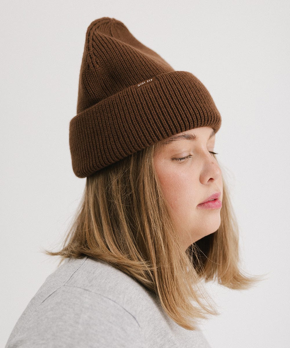 Woman wearing a chocolate colored oversized knit beanie, facing sideways against a plain background #color_chocolate
