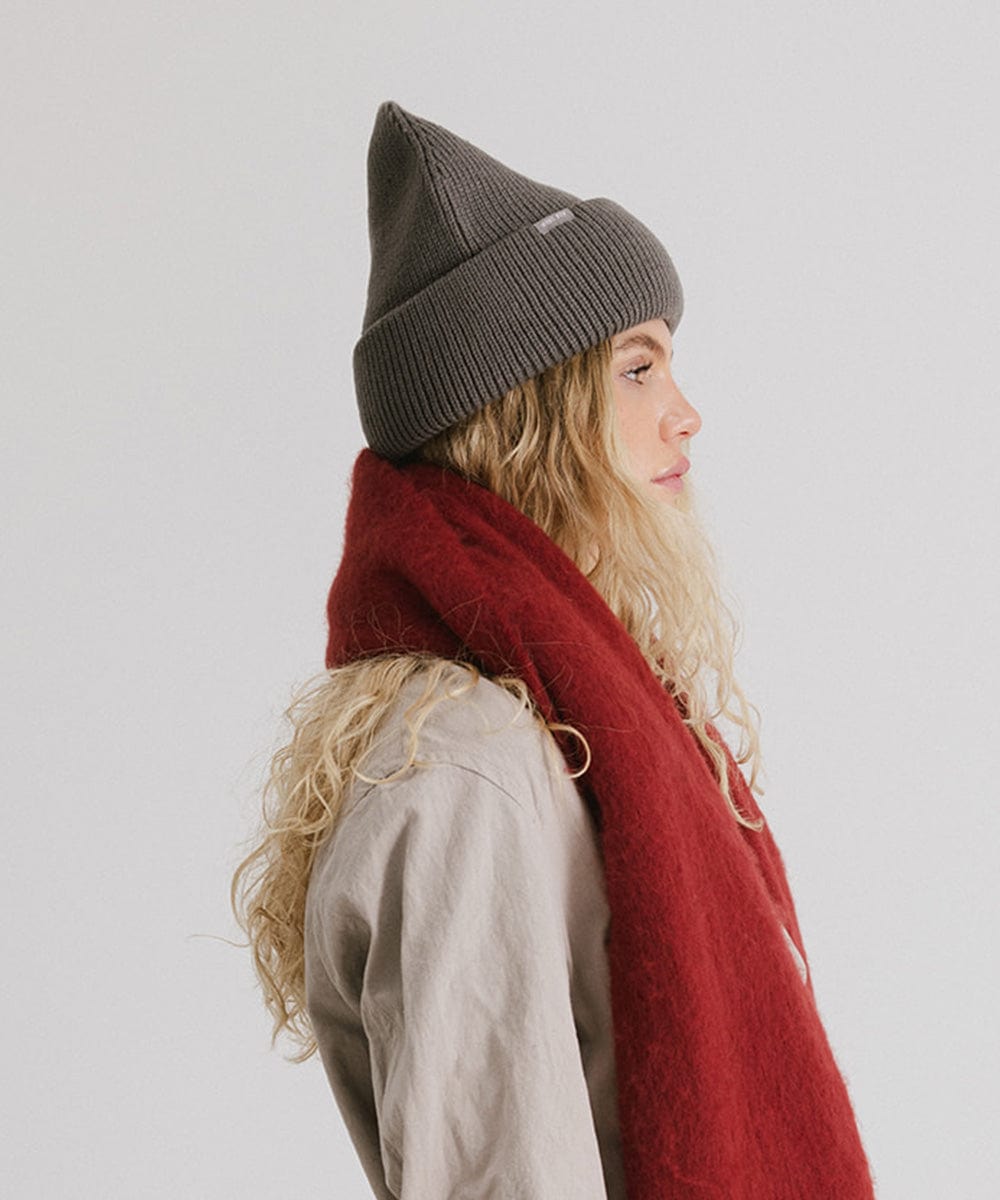 Woman wearing a grey oversized knit beanie, a cream coat and a red oversized scarf, facing sideways against a plain background #color_grey