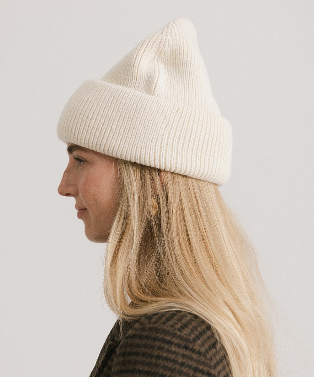 Woman wearing an off white oversized knit beanie, facing sideways against a plain background #color_off white