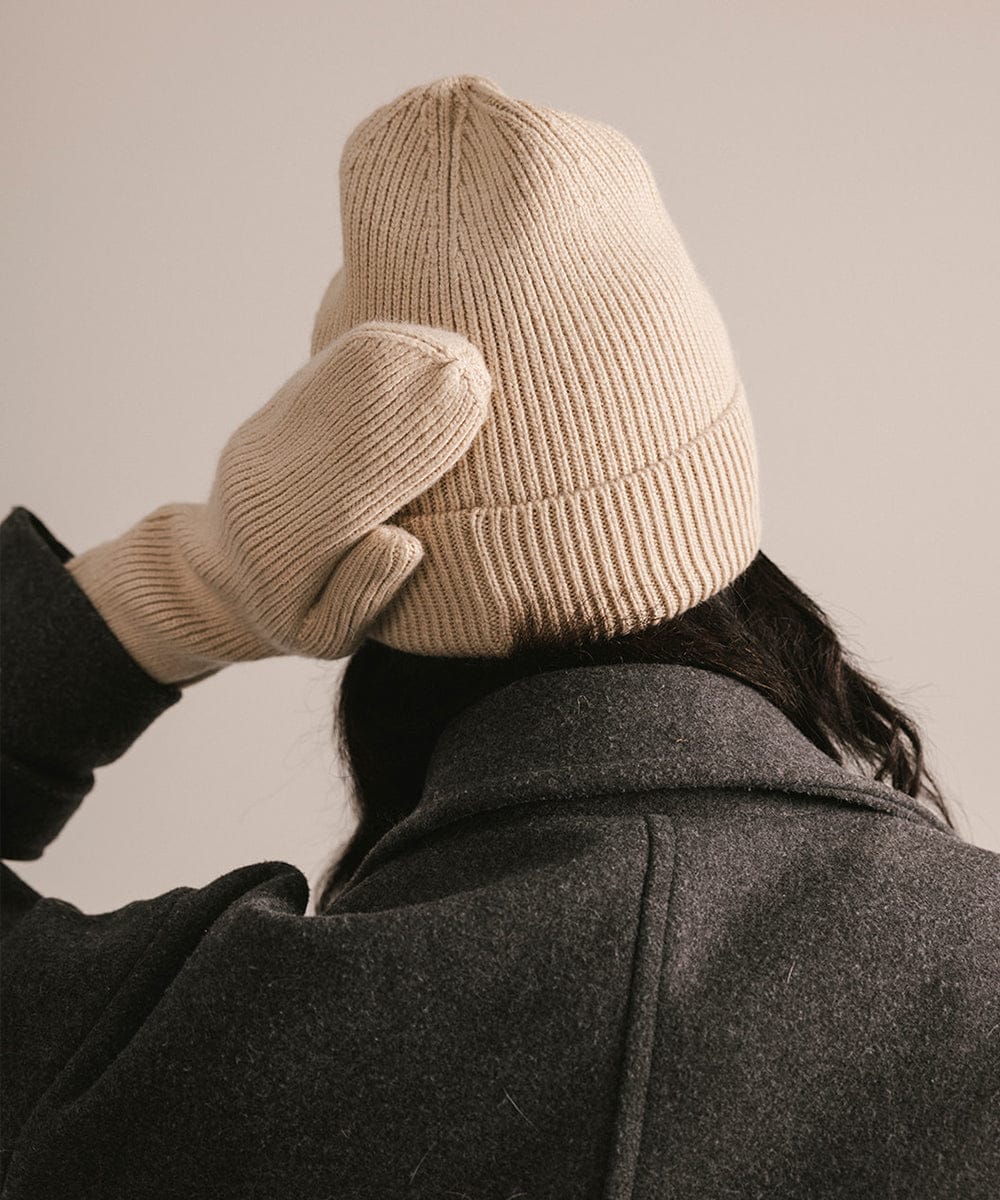 Person wearing a beige knit beanie and gloves, adjusting the beanie against a neutral background. #color_taupe
