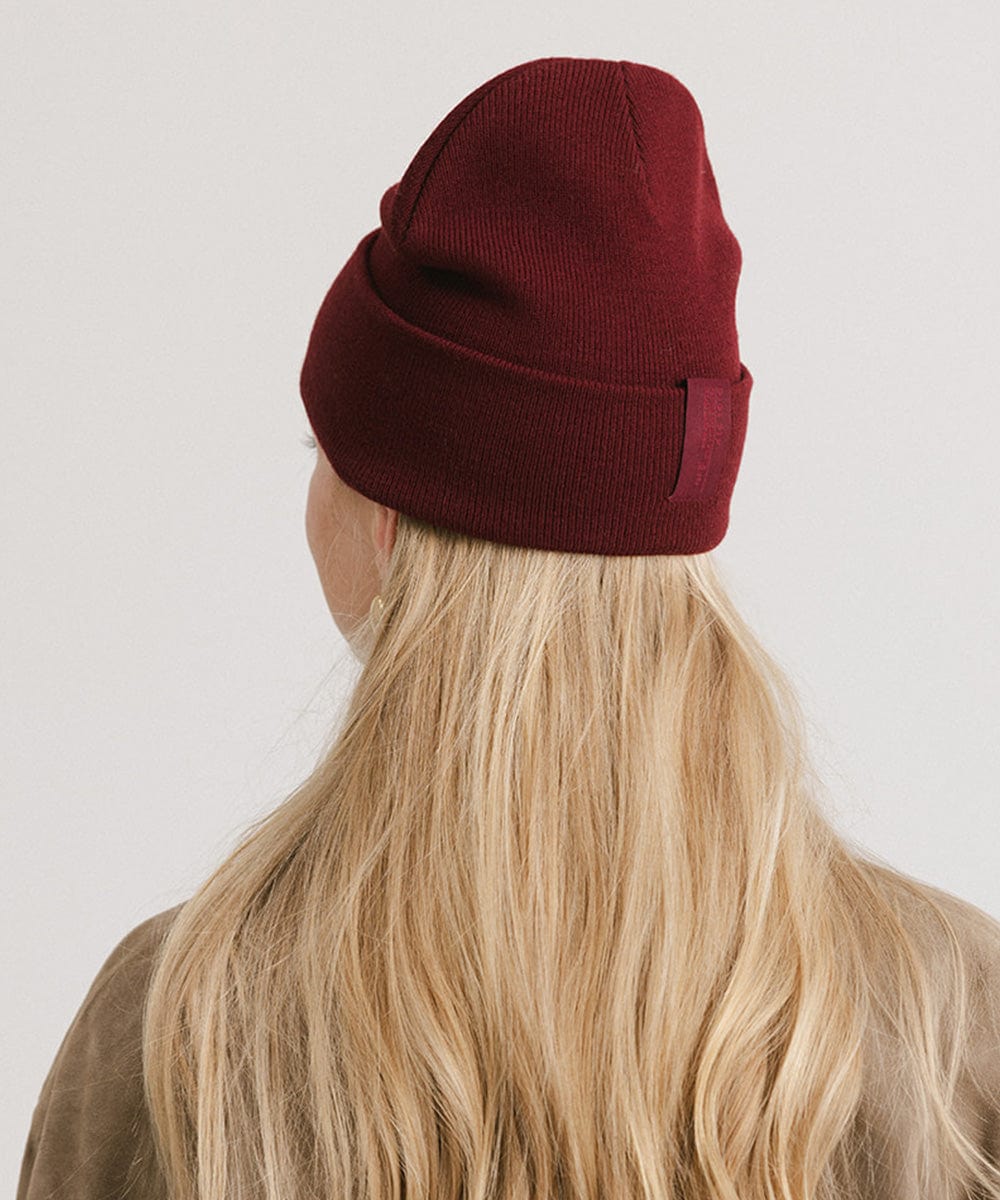 Woman with blonde hair wearing a burgundy beanie and a beige shirt, facing behind against a plain background #color_burgundy
