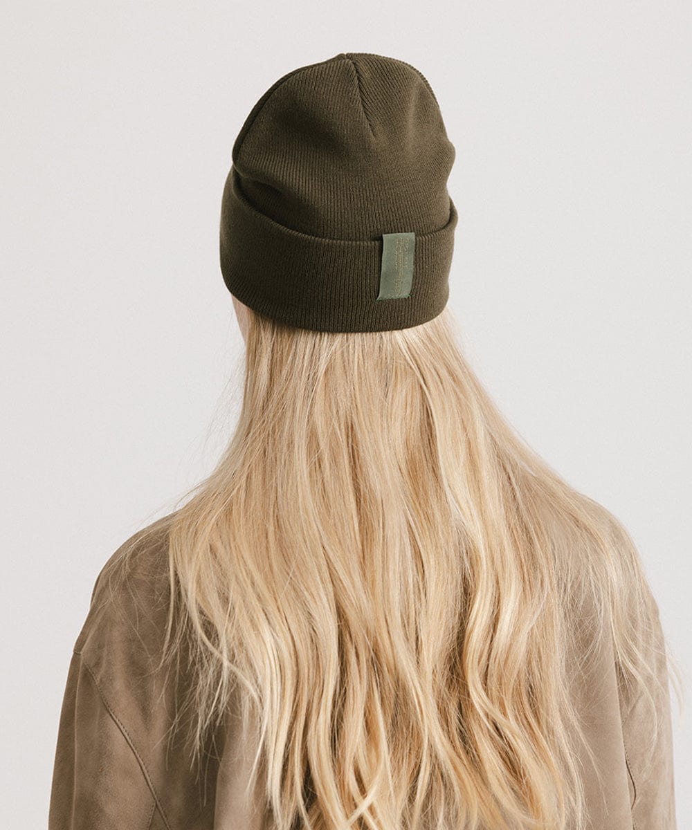 Woman with blonde hair wearing a dark green beanie and a beige shirt, facing behind against a plain background #color_dark green