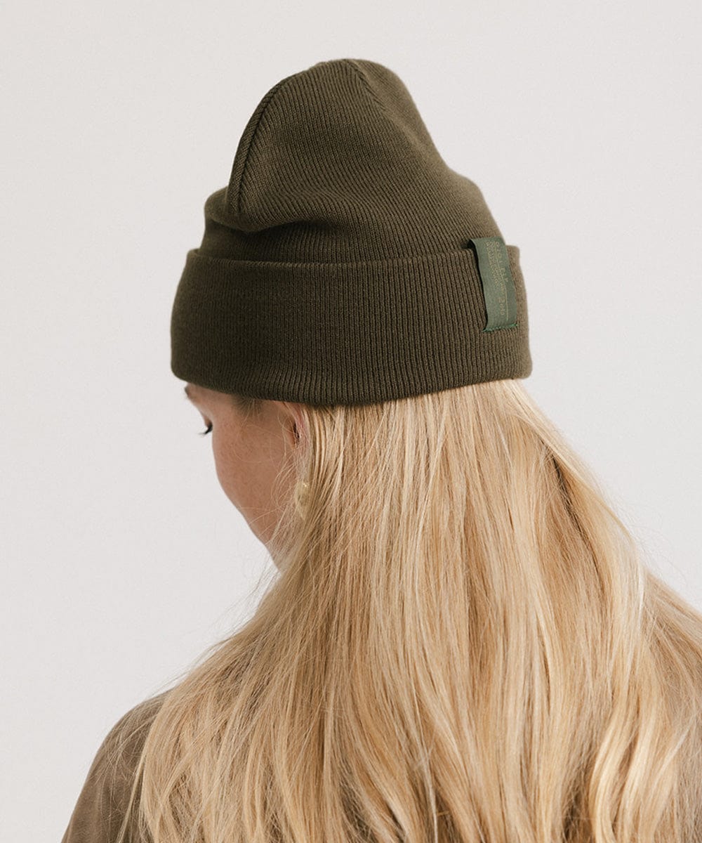 Woman with blonde hair wearing a dark green beanie facing behind against a plain background #color_dark green