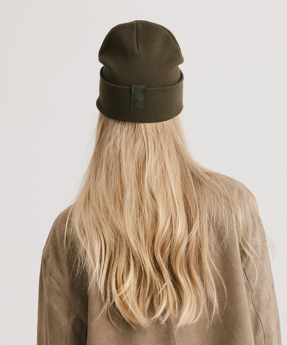 Woman with blonde hair wearing a dark green beanie and a beige shirt, facing behind against a plain background #color_dark green