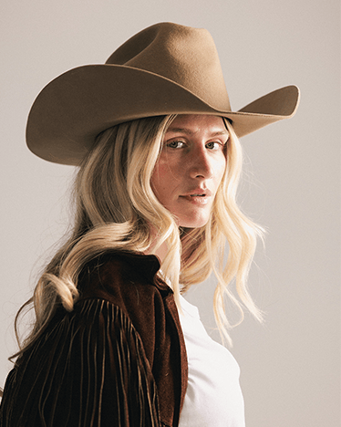 Woman wearing a brown cowboy hat and dark jacket against a plain background