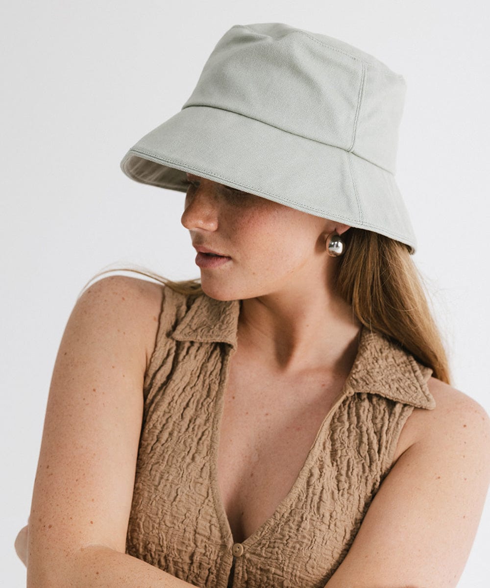Woman wearing a sage packable bucket hat and a brown top, posing sideways against a plain background  #color_sage