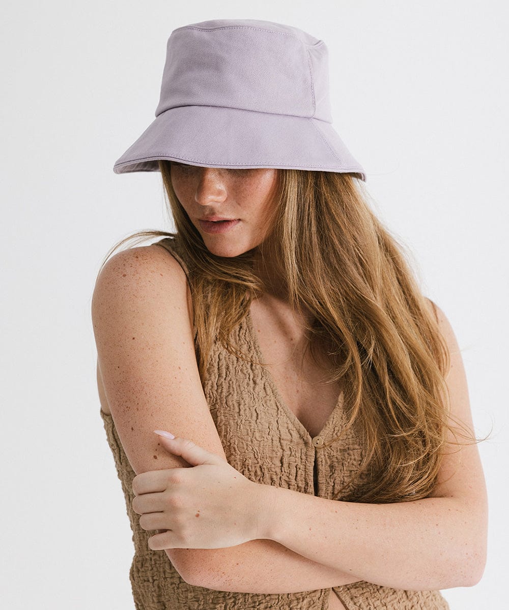 Woman wearing a lavender packable bucket hat and a brown top, posing  against a plain background #color_lavender