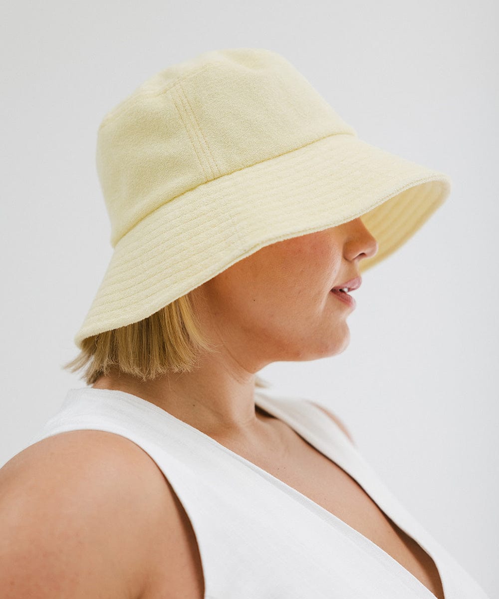 Woman wearing a butter yellow colored bucket hat and a white top, facing sideways against a plain background #color_butter yellow