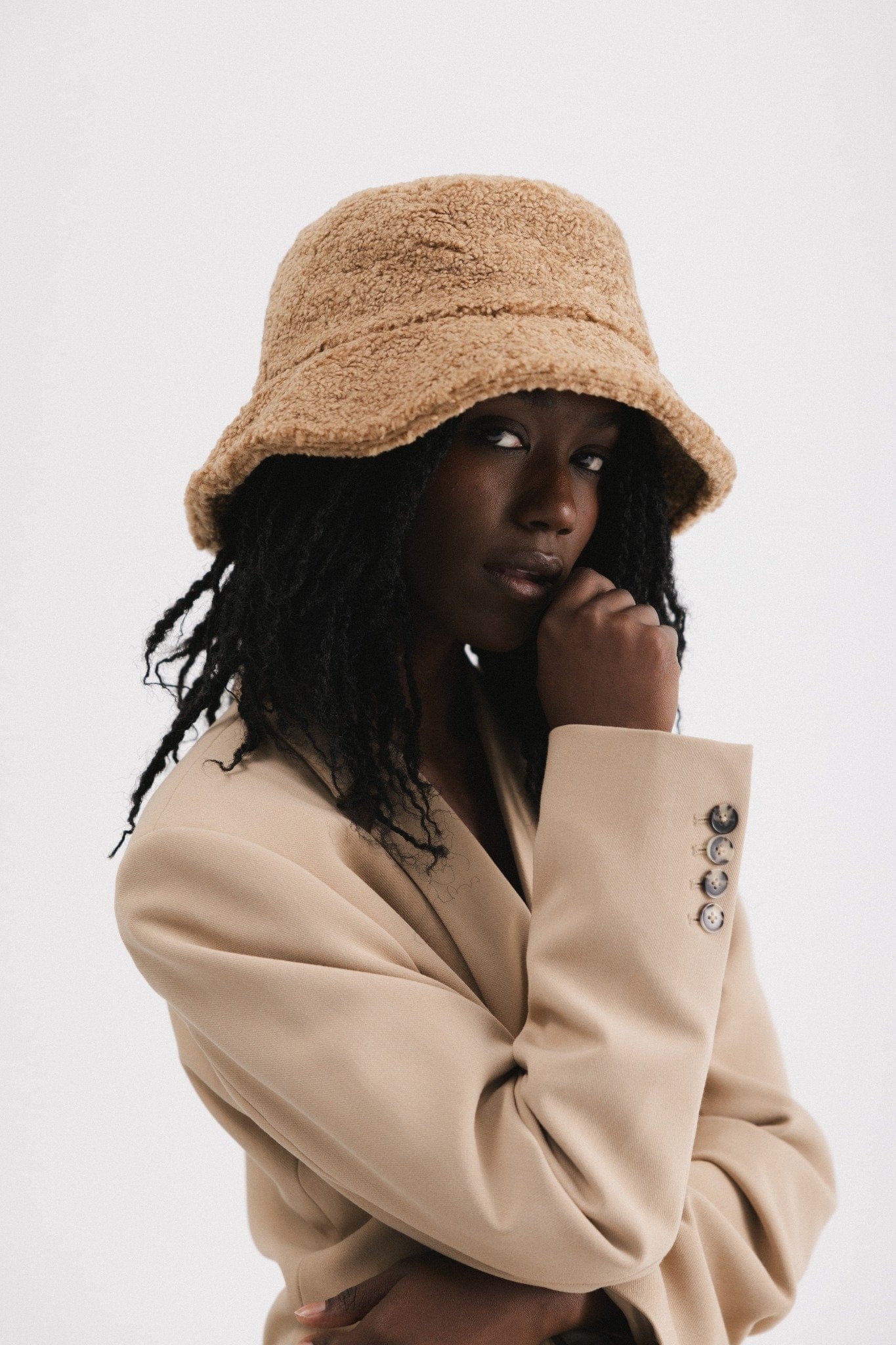 Woman wearing a camel colored sherpa bucket hat and a beige coat, facing sideways against a plain background #color_camel