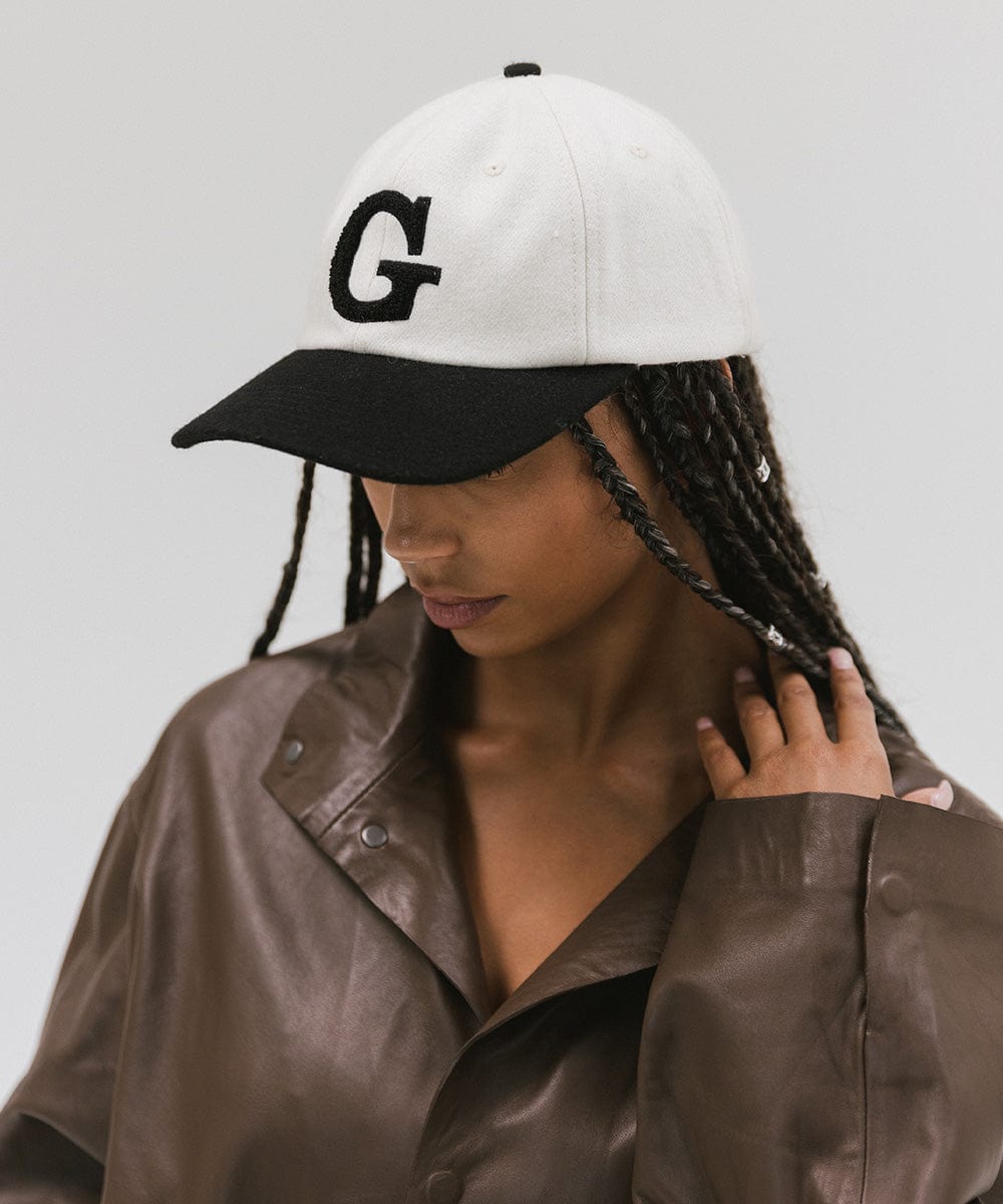 Woman wearing a cream and black baseball cap with the letter 'G' and a brown jacket, posing against a plain background #color_cream-black
