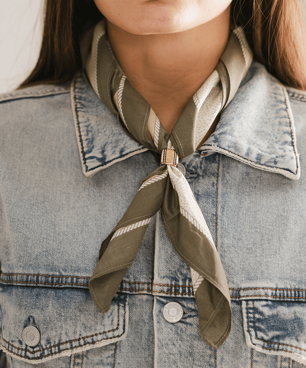 Person wearing a green and beige striped scarf and Aztec Square Concho Bandana Slide around their neck with a denim jacket. #color_gold