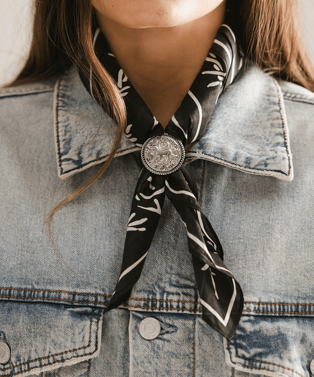 Person wearing a black and white patterned scarf with a Horse Concho Bandana Slide on a denim jacket. #color_silver