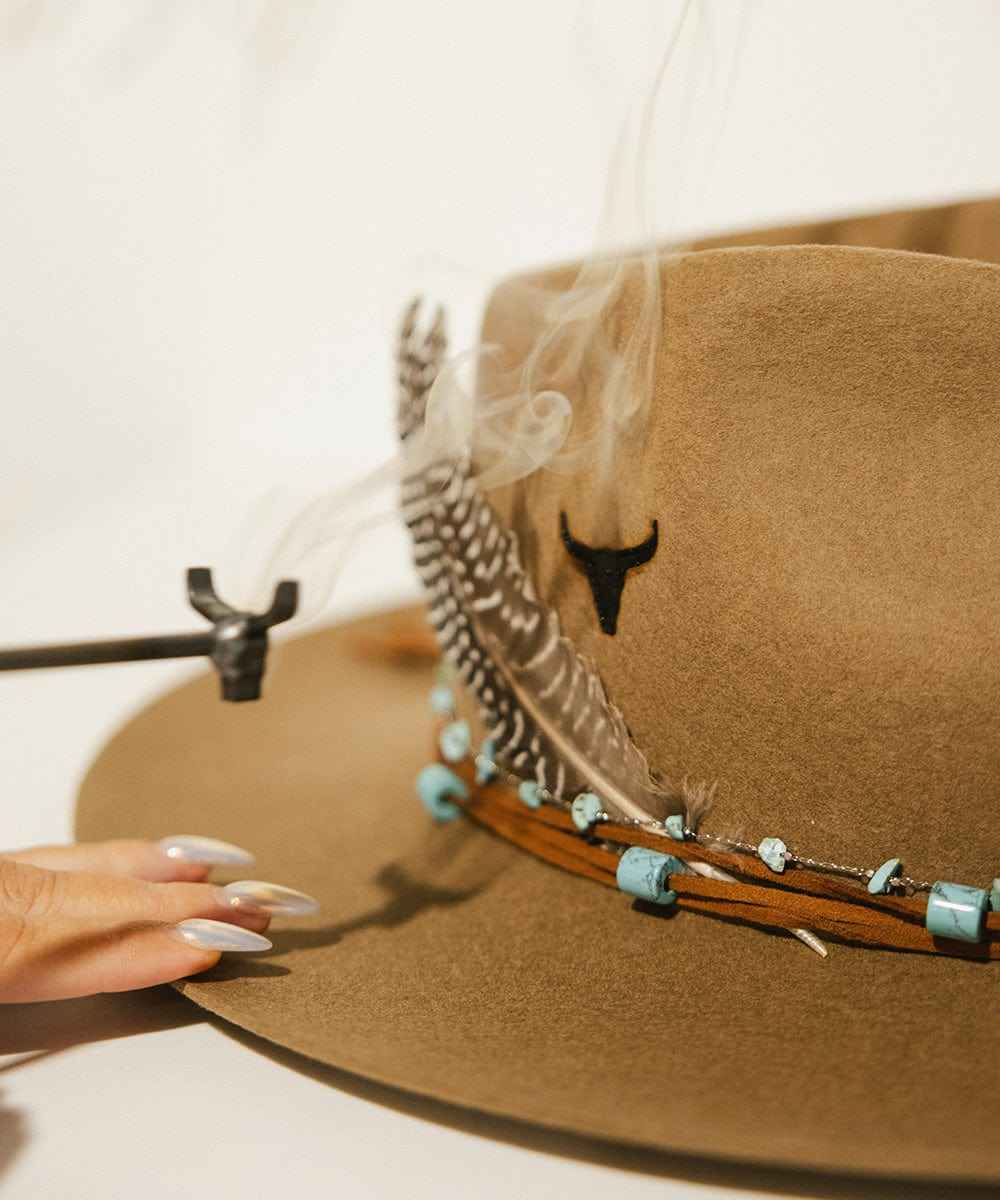 Brown hat with feather and decorative band held by a hand against a white background