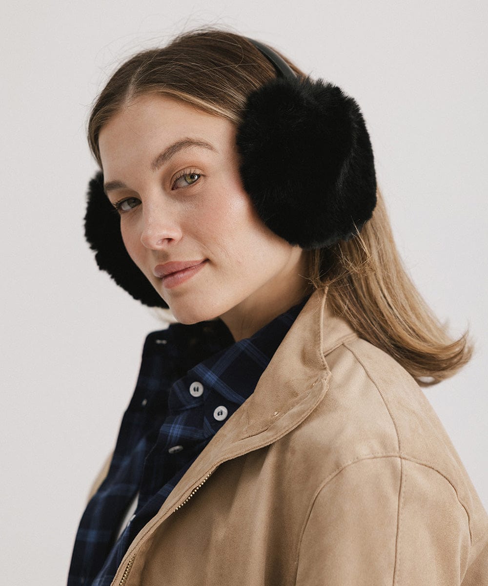 Woman wearing black fur ear muffs and a beige jacket, posing sideways against a plain background #color_black