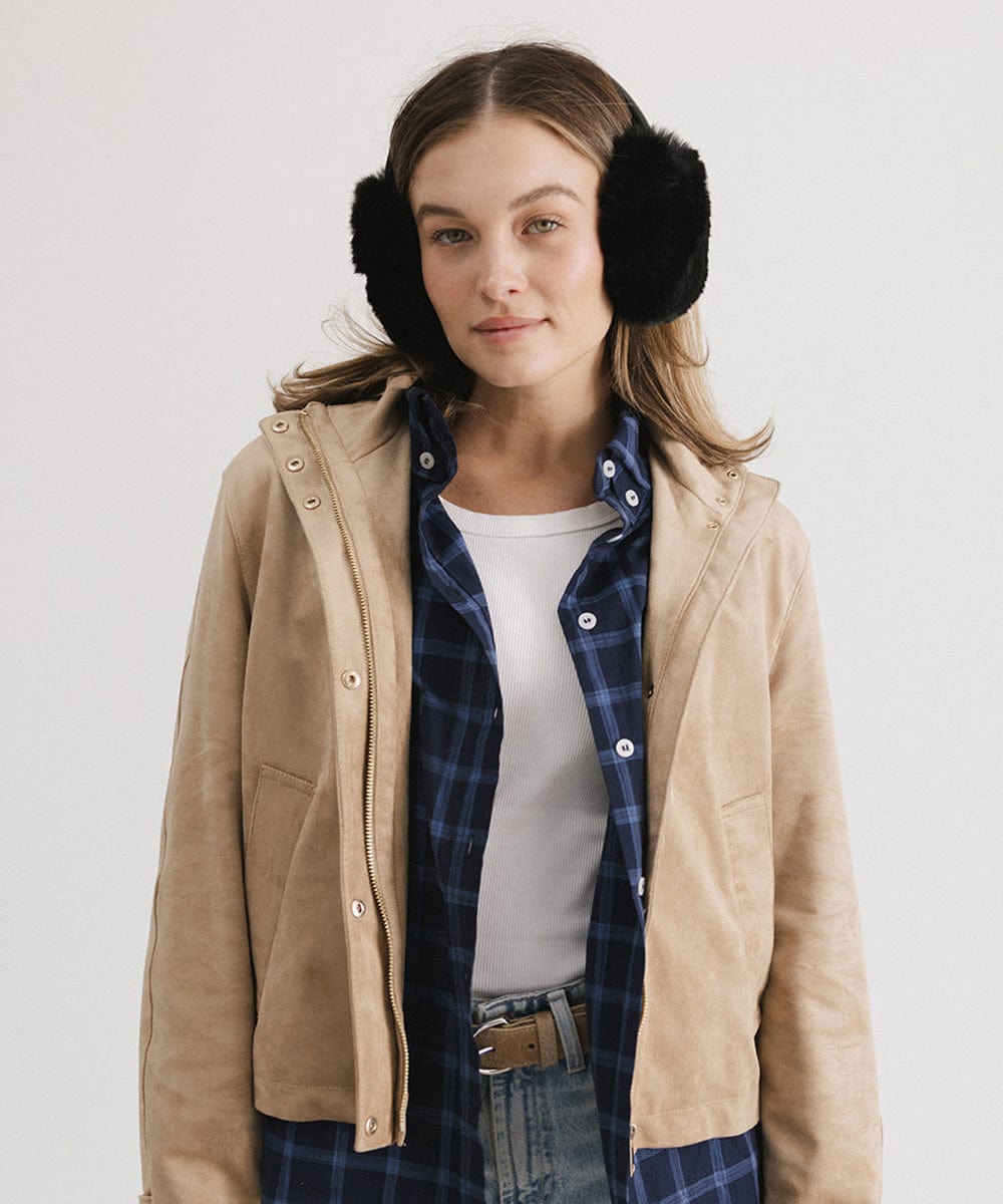 Woman wearing black fur ear muffs and a white top with a beige jacket against a plain background #color_black