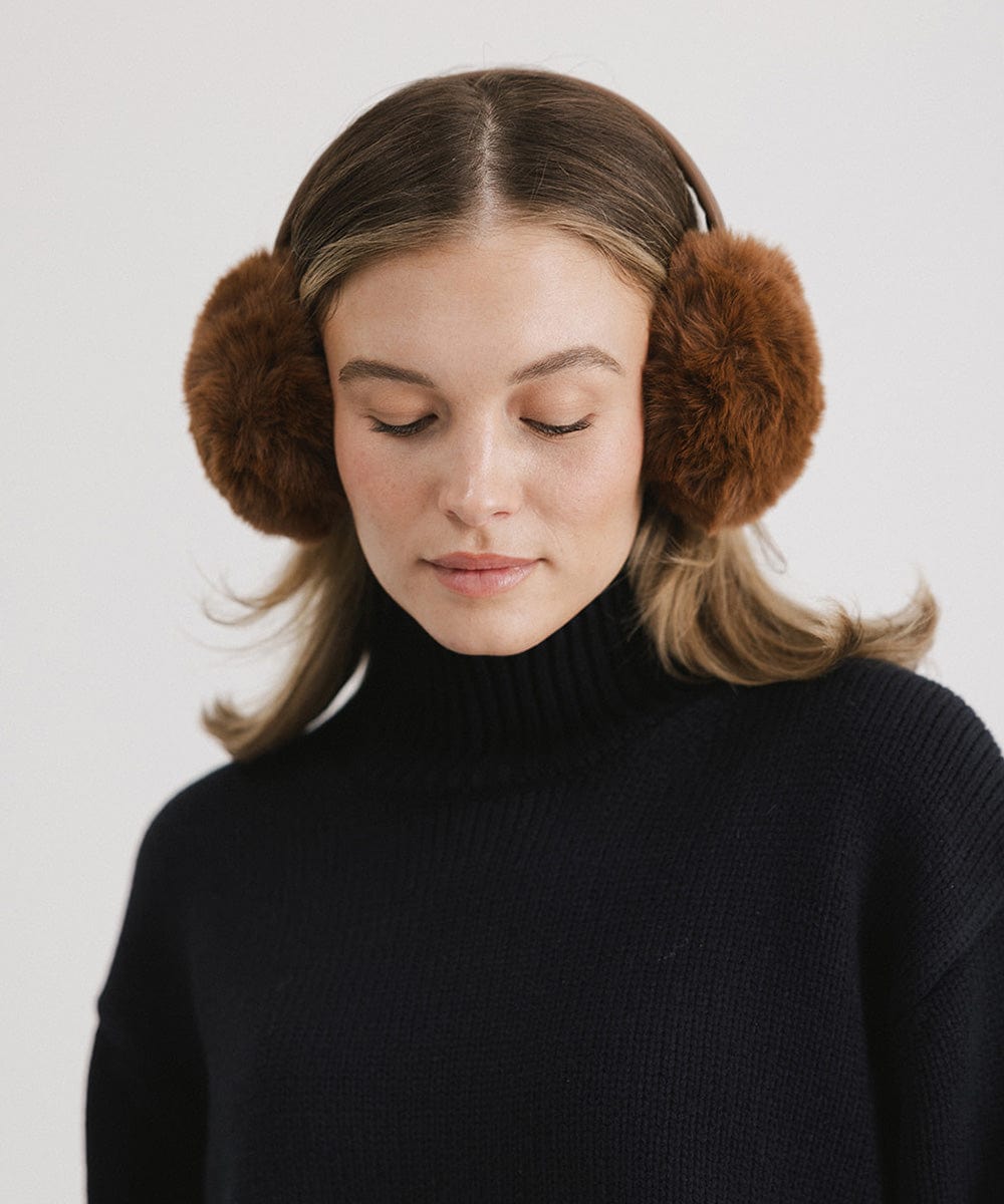 Woman wearing brown fur ear muffs and a black sweater, looking down against a plain background #color_brown