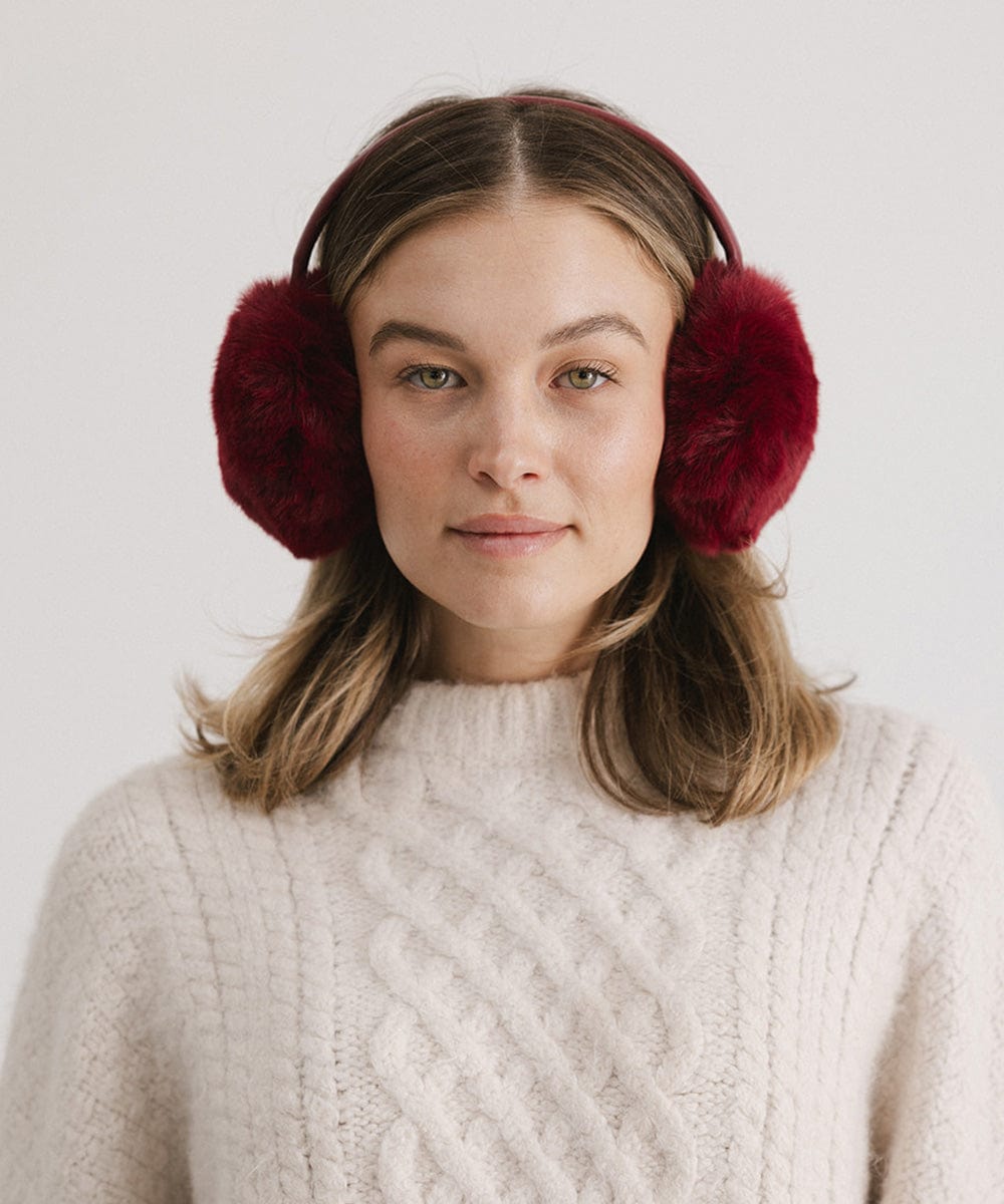 Woman wearing burgundy fur ear muffs and a white sweater against a plain background #color_burgundy