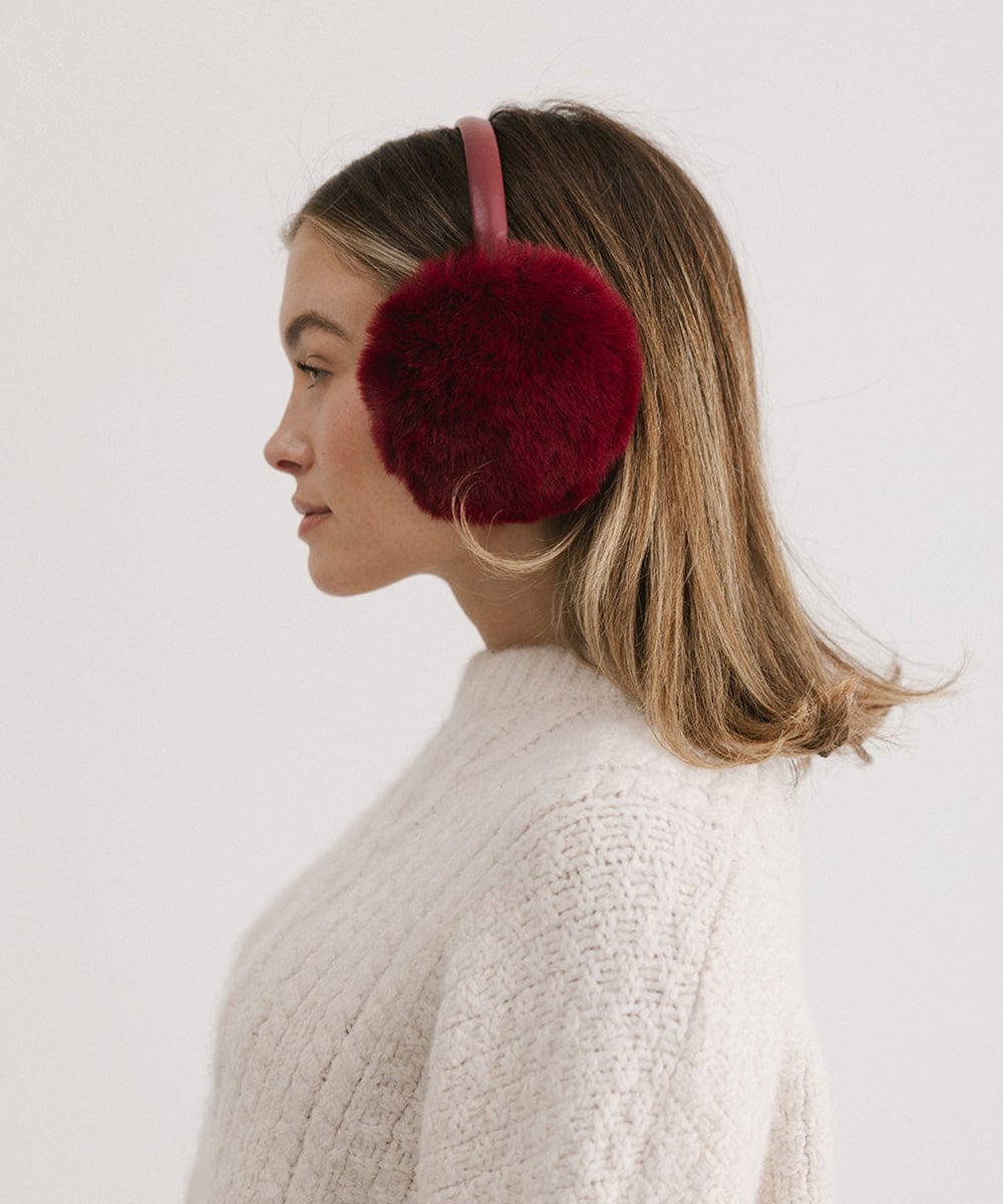Woman wearing burgundy fur ear muffs and a white sweater, facing sideways against a plain background #color_burgundy