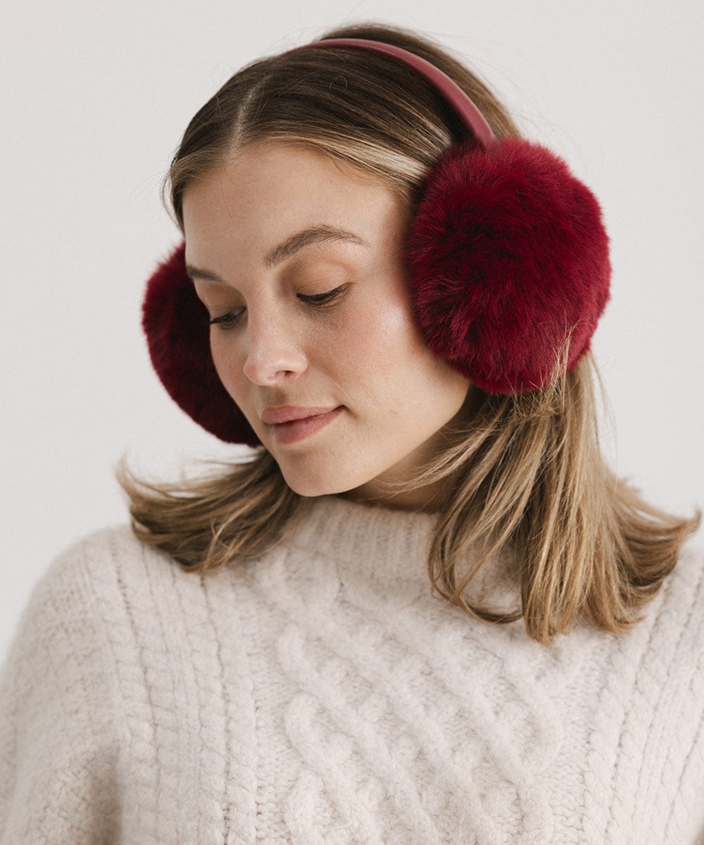 Woman wearing burgundy fur ear muffs and a white sweater, posing against a plain background #color_burgundy