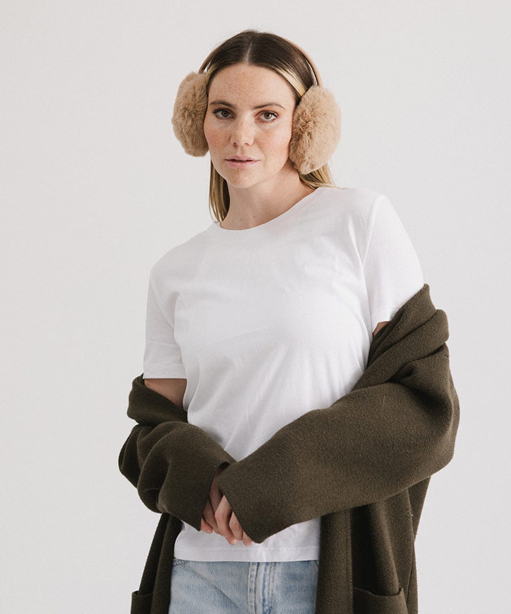 Woman wearing tan fur ear muffs and a white t-shirt with a brown coat against a plain background #color_tan