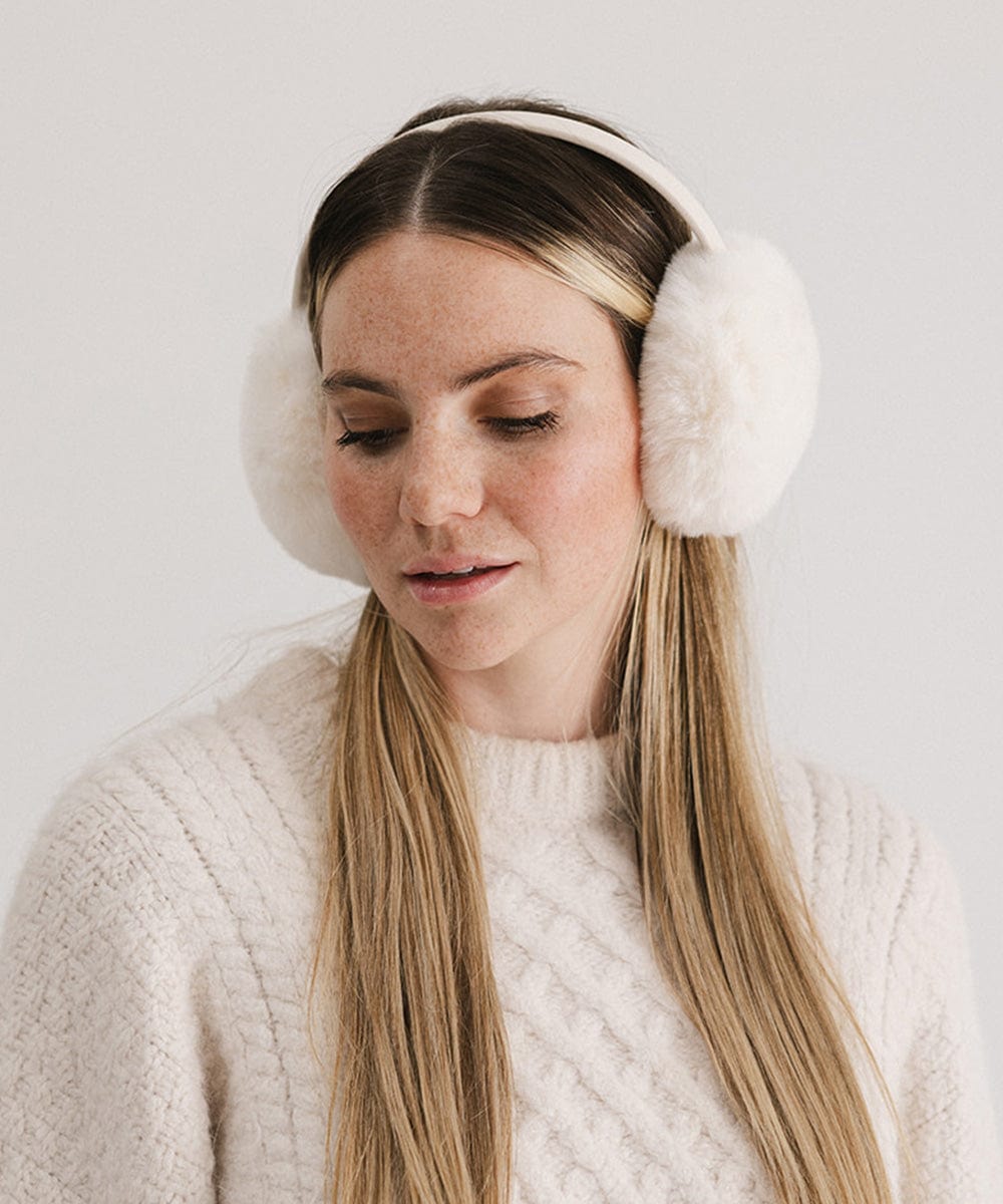 Woman wearing winter white colored fur ear muffs and a white sweater, posing against a plain background #color_winter white