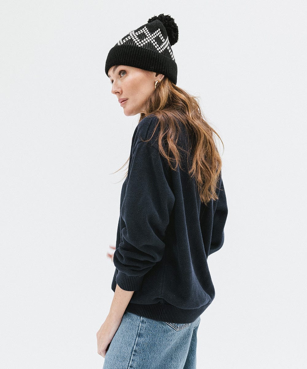 Woman wearing a black knit beanie with a pom-pom, a dark blue sweater and blue jeans, posing sideways against a plain background #color_black