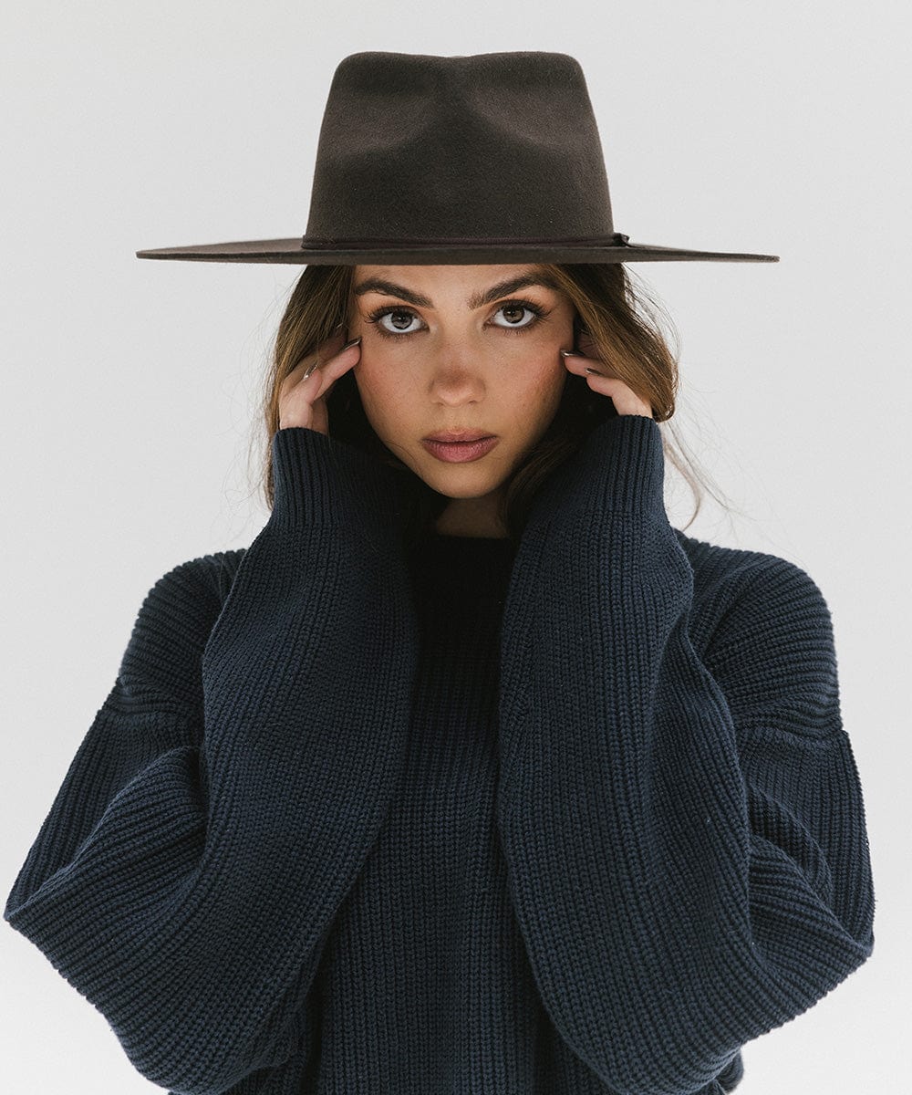 Woman wearing a dark brown Emery Teardrop Fedora Hat and a dark blue sweatshirt against a plain background #color_dark brown