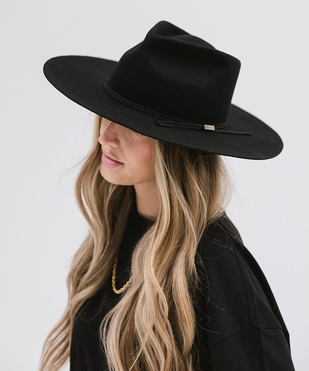 Woman with long hair wearing a black Emery Teardrop Fedora Hat with a hat band and a black t-shirt against a plain background #color_black