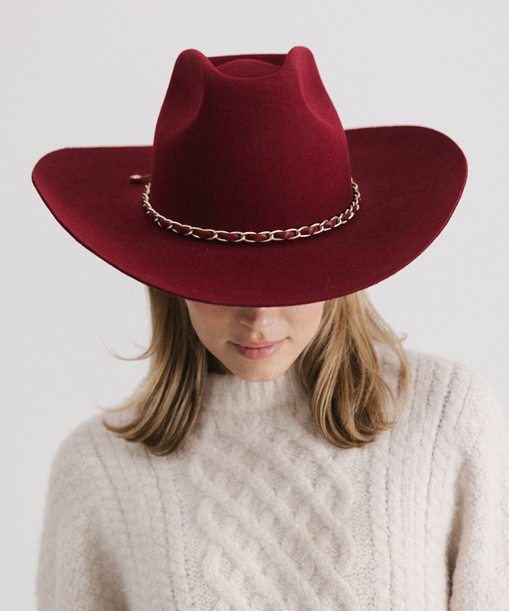 Person wearing a burgundy colored felt hat with a metallic chain detail on the band and a white sweater against a plain background   #color_burgundy