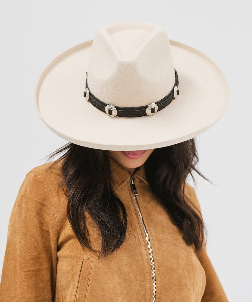 Woman wearing an off white colored pencil brim fedora hat with a wide brim and a hat band, and a brown jacket against a white background  #color_off white