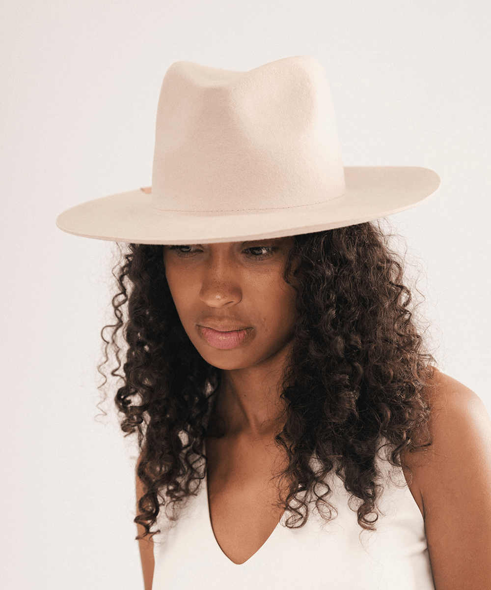 A woman with long, curly dark hair wearing a light beige wide-brim fedora hat and a white sleeveless top, photographed against a plain light background #color_cream