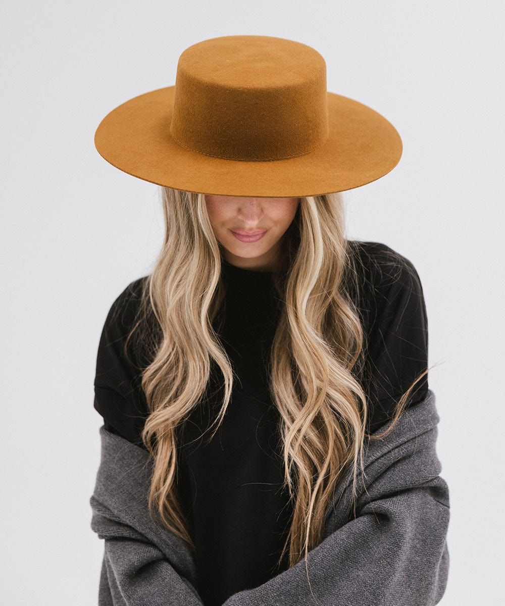 Woman wearing a brown boater-style hat, a black t-shirt and grey jacket, looking down against a plain background #color_brown