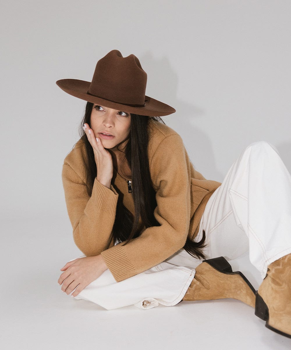 Woman wearing a dark oak colored Western hat with a classic cattleman crown, a brown jacket, white pants and brown boots against a plain background  #color_dark oak