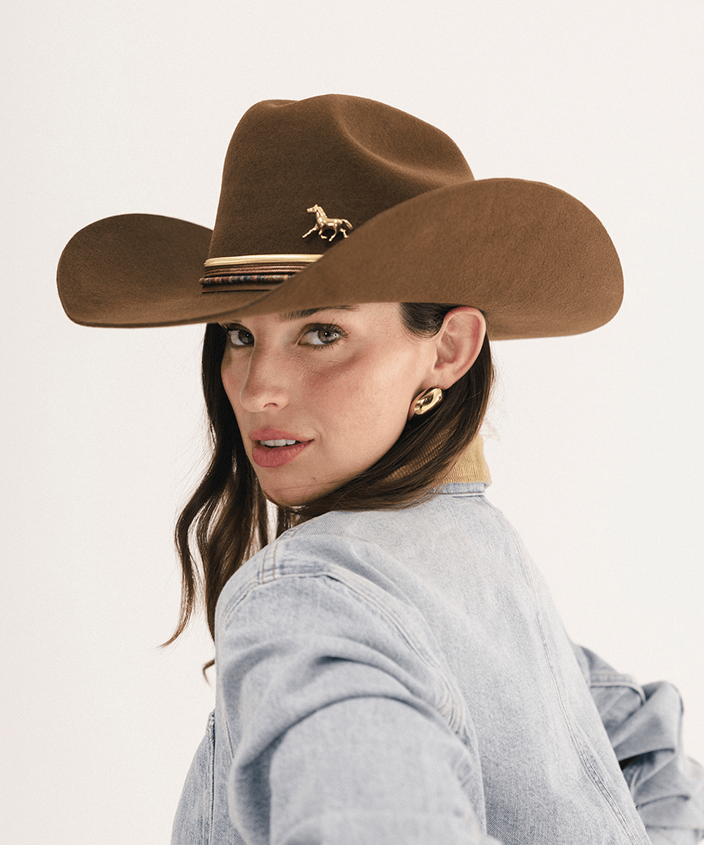 A woman wearing a brown cowboy hat with a small horse emblem and striped band, paired with a light blue denim shirt against a plain white background #color_coffee