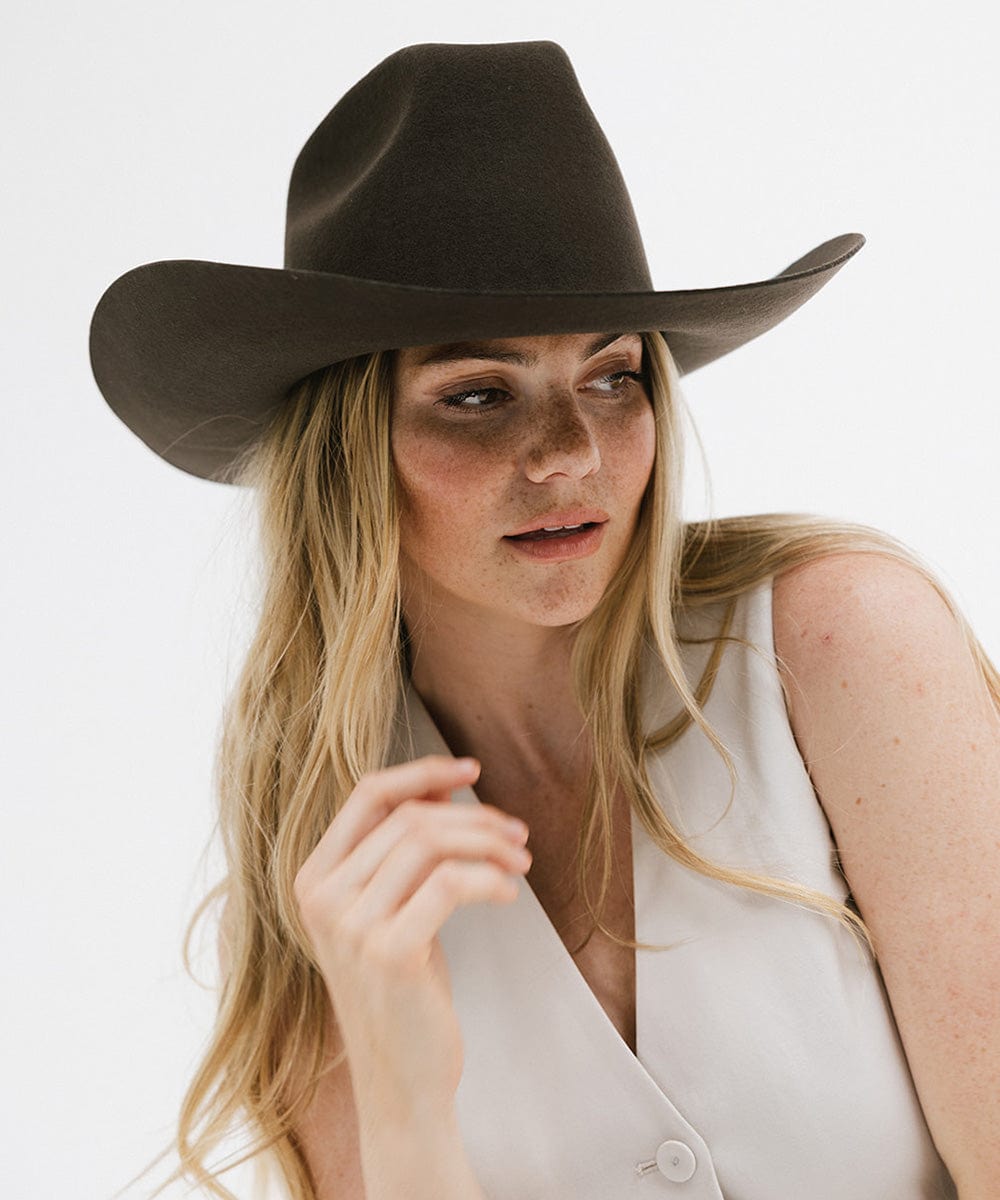 Woman wearing a dark brown cowboy hat and a white sleeveless jacket against a plain background #color_dark brown