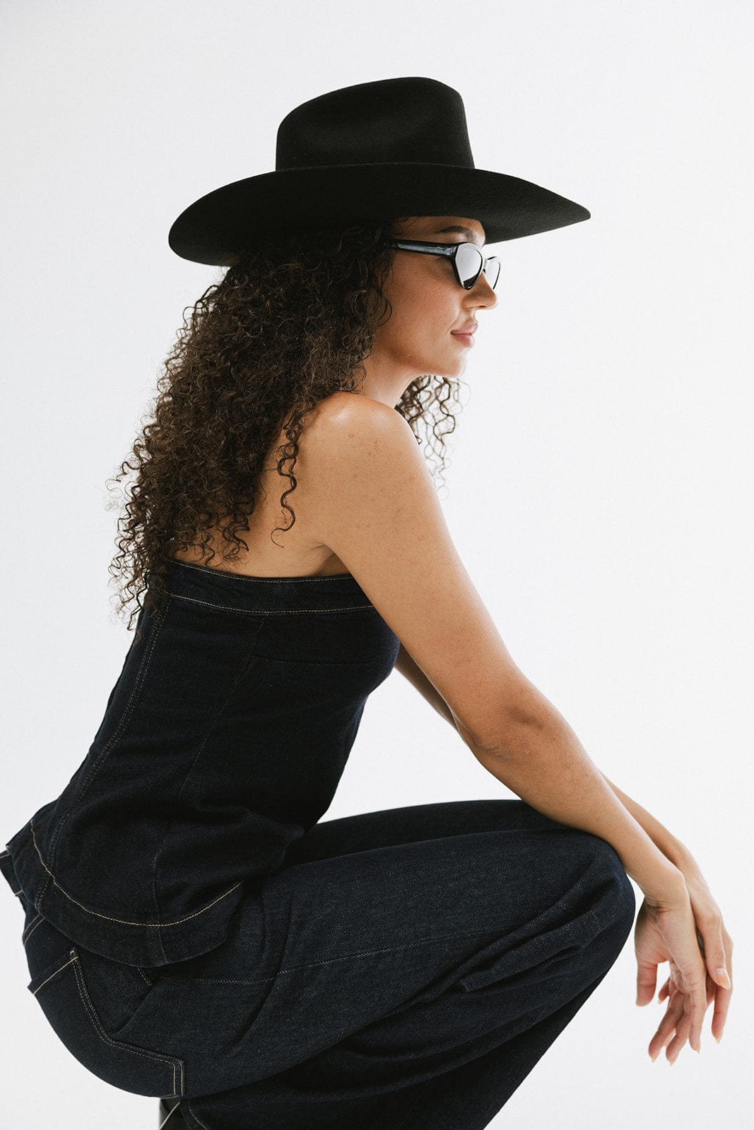 Woman wearing a black cowboy hat, sunglasses, a dark blue denim top and dark blue denim jeans a plain background #color_black
