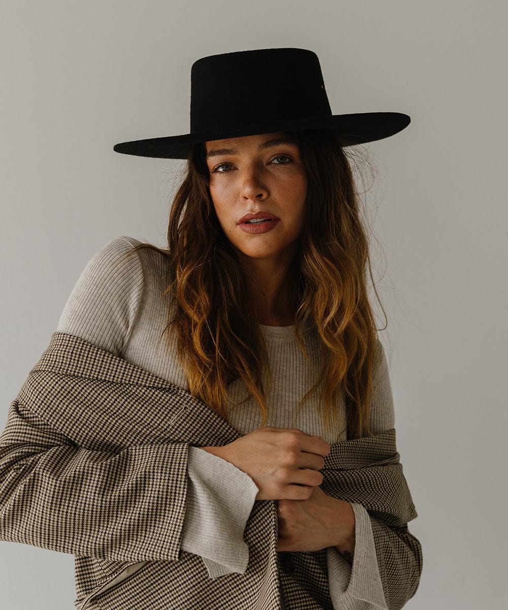 Woman wearing a black boater hat and a white top with a coat against a plain background #color_black