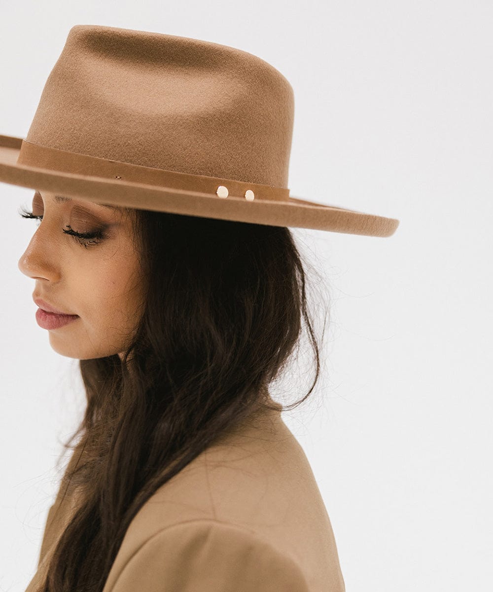 Woman wearing a tan felt fedora hat with a hat band and a brown coat, facing sideways against a plain background #color_tan