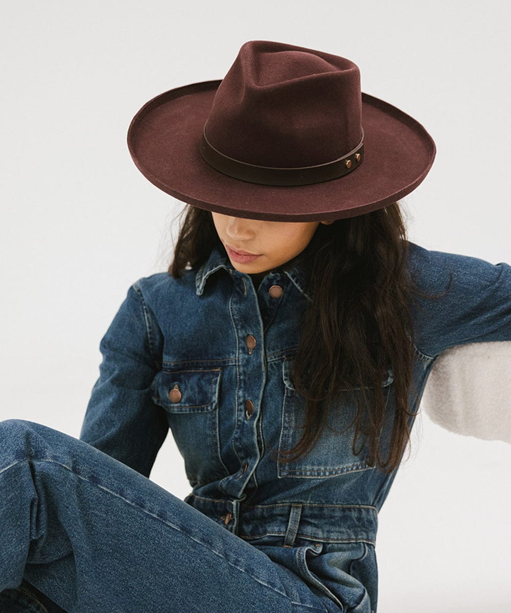 Woman wearing a dark cherry colored felt fedora hat with a hat band, a denim shirt and jeans, sitting against a plain background #color_dark cherry