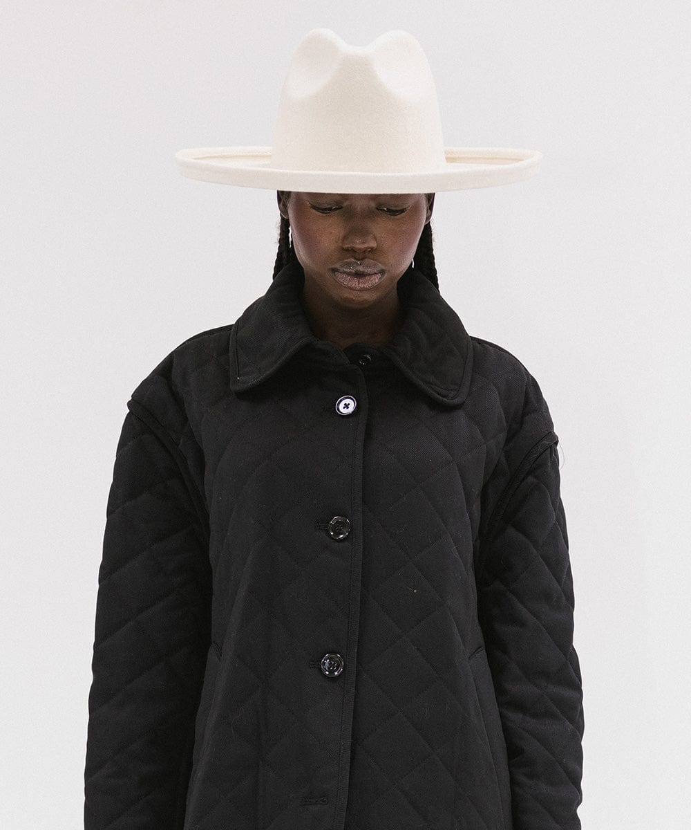 Person wearing an off white wide brim felt fedora hat and a black coat against a white background #color_off white
