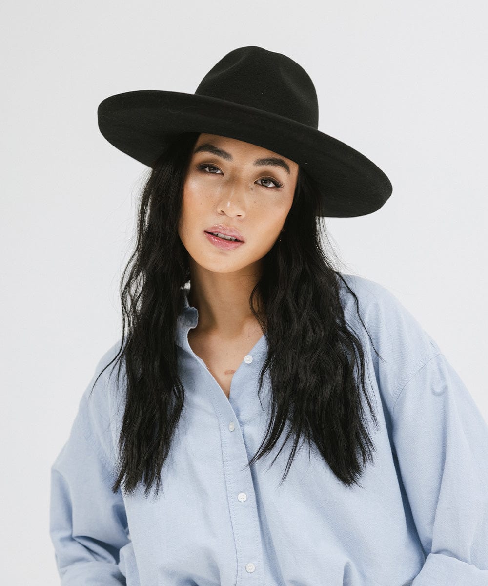 Woman wearing a black wide brim felt fedora hat and a blue shirt against a white background #color_black