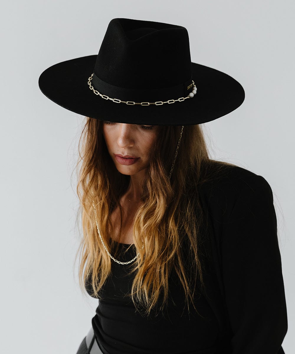 Woman wearing a black wide brim fedora hat with a gold chain detail around the base of the crown and a black top against a plain background  #color_black