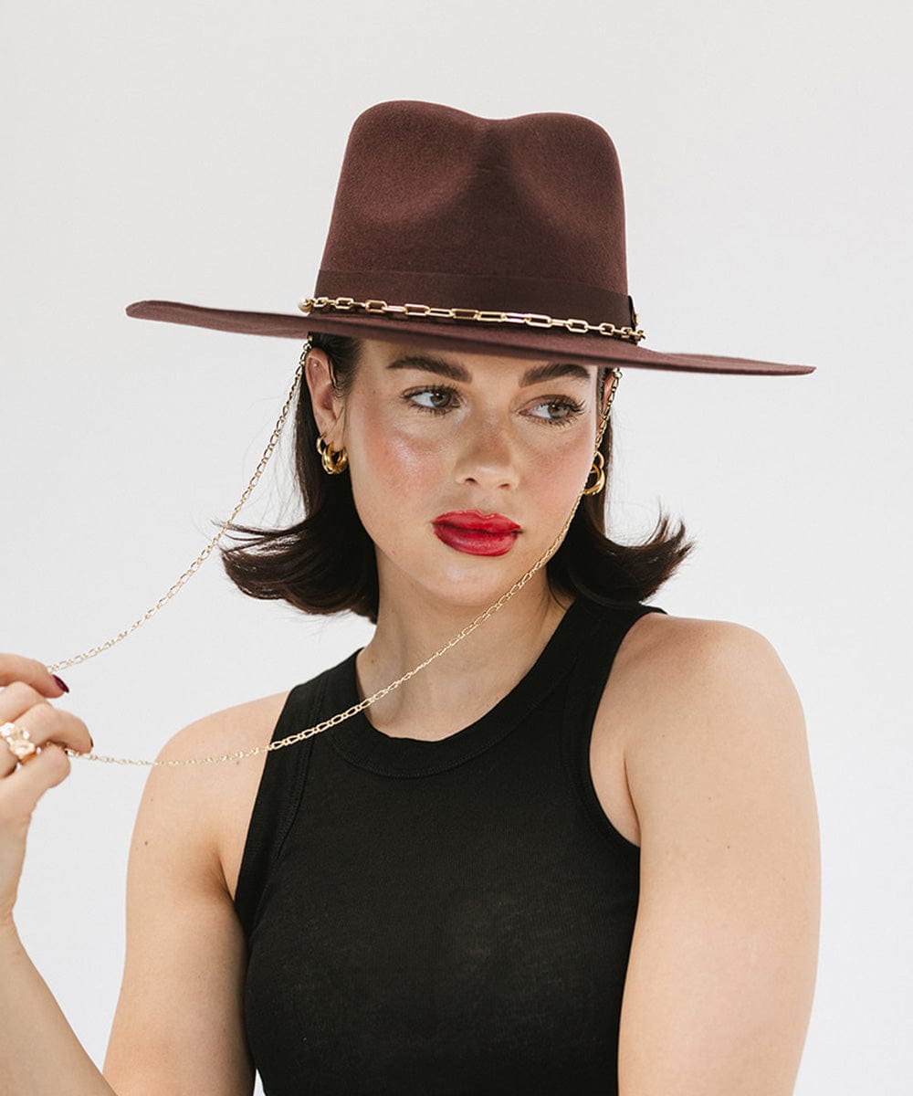 Woman wearing a dark cherry colored wide brim fedora hat with a gold chain detail around the base of the crown and a black top against a plain background #color_dark cherry