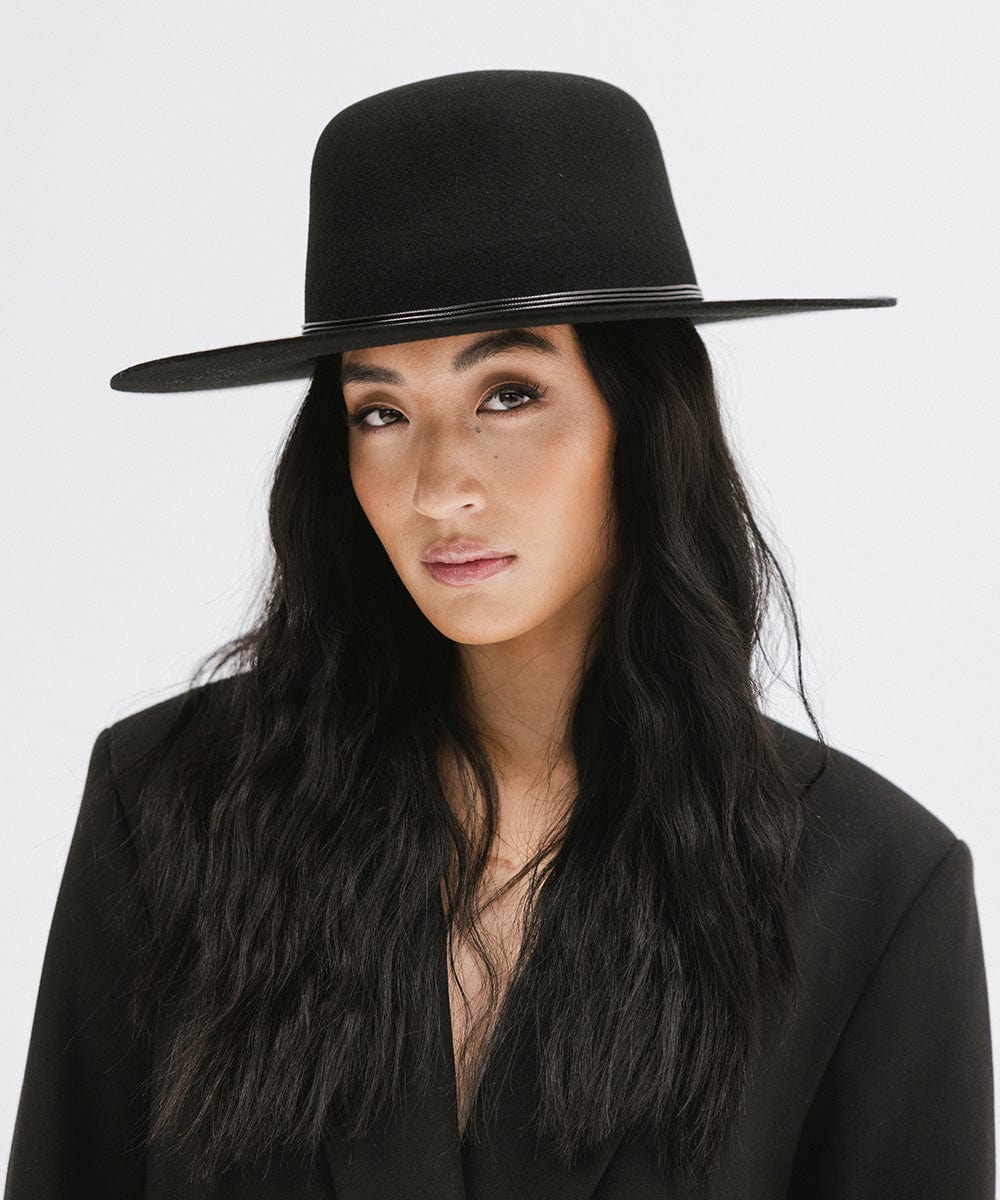 Woman wearing a black open crown hat with a hat band and a black blazer against a plain background #color_black